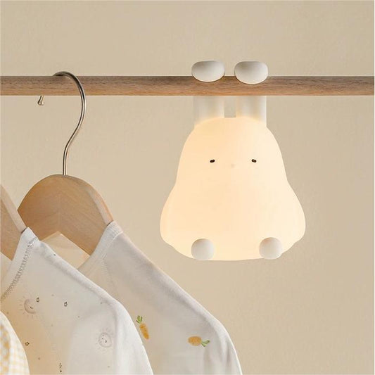 A foldable ear bunny-shaped silicone pat night light hanging from a wooden hanger, emitting a warm white light.
